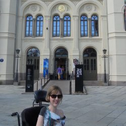 Woman in a wheelchair smiles in front of a large Oslo museum building with banners reading “Aung San Suu Kyi” and “Mother Democracy.”