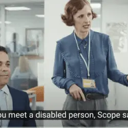 A woman in office attire with a staff ID badge gestures animatedly while standing next to a man in a suit who is seated and smiling slightly. The setting is a modern office with desks and glass partitions in the background. Subtitles at the bottom read, "Should you meet a disabled person, Scope says hide."