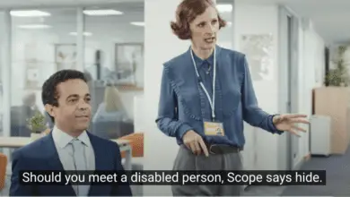 A woman in office attire with a staff ID badge gestures animatedly while standing next to a man in a suit who is seated and smiling slightly. The setting is a modern office with desks and glass partitions in the background. Subtitles at the bottom read, "Should you meet a disabled person, Scope says hide."