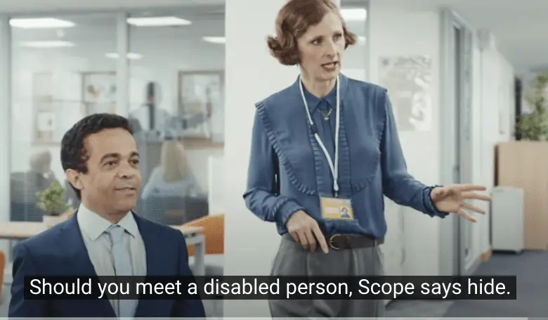 A woman in office attire with a staff ID badge gestures animatedly while standing next to a man in a suit who is seated and smiling slightly. The setting is a modern office with desks and glass partitions in the background. Subtitles at the bottom read, "Should you meet a disabled person, Scope says hide."