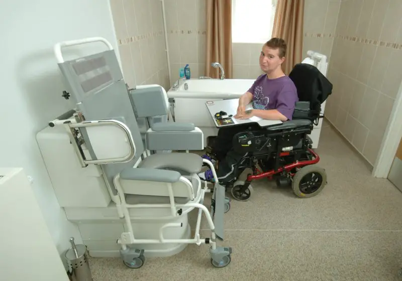 Teen boy in a powered chair with a fixed plastic tray in a bathroom with a toilet chair adaption