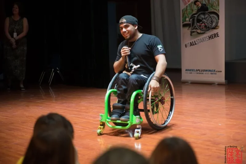 Adam Fotheringham at speaking at an event he is in a green sports wheelchair and has a baseball cap on.