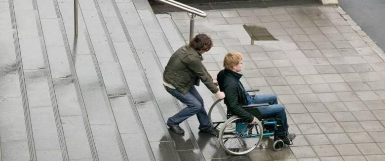 Man struggling to help disabled man in wheelchair up stairs