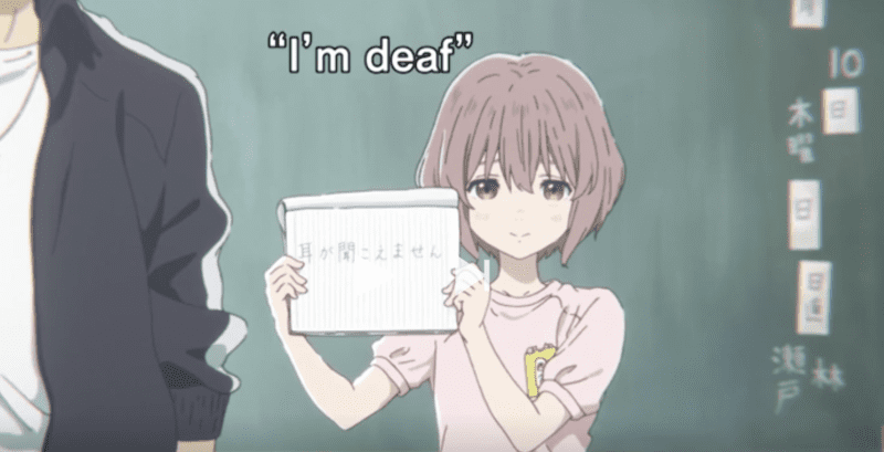Anime girl in classroom holding up a paper book with the words "I'm Deaf" on it to show new classmates. Screenshot from A Silent Voice The Movie