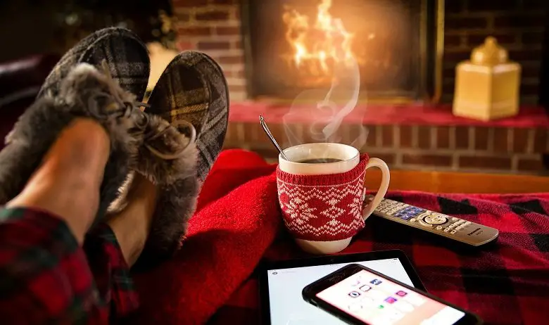 Legs in slippers resting on a footstall with a phone, TV remote and tablet on in front of a fire