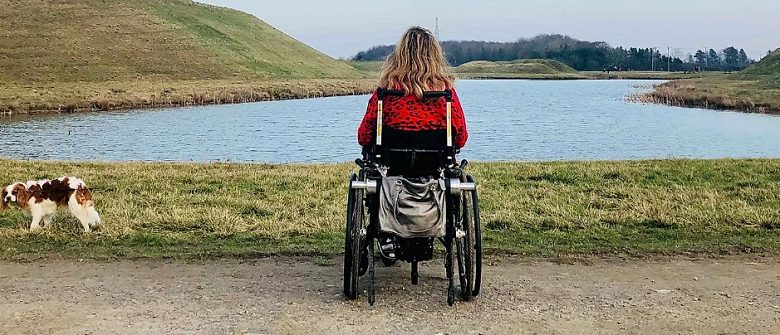 Carrie-Ann in her wheelchair in front of a lake with her dog