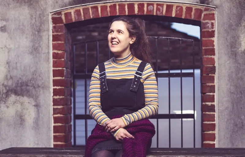 Disabled comedian Rosie Jones sat on a bench in front of a brick wall and window