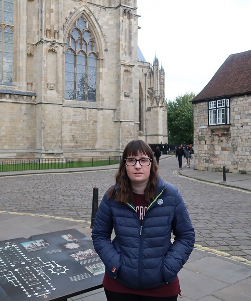 Holly stood in front of York Minster, cobbled street and houses in background, wearing blue jacket