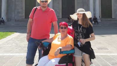 Caroline in her wheelchair with pink hair and a yellow t-shirt with her two children in front of a historic building