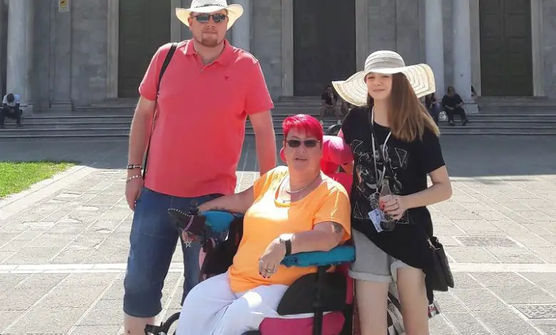 Caroline in her wheelchair with pink hair and a yellow t-shirt with her two children in front of a historic building