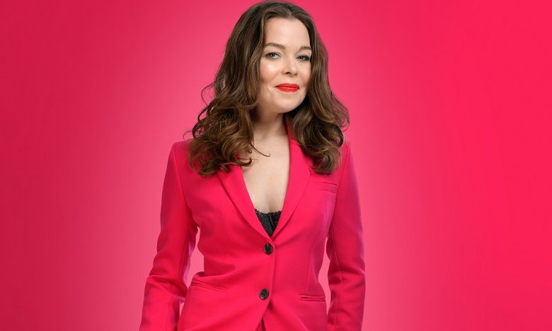 Juliette Burton in a pink suit and white heeled shoes stood on a pink background