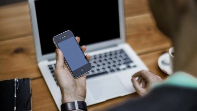 Man holding a mobile phone sat at a desk with his laptop