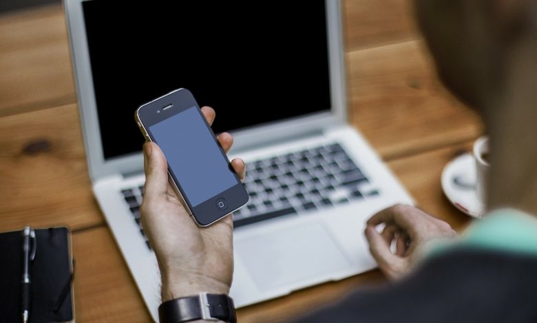 Man holding a mobile phone sat at a desk with his laptop