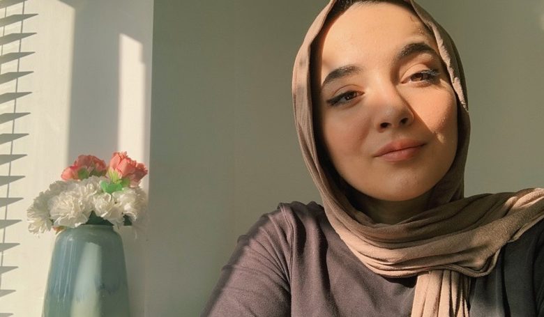 Lina Bettayeb wearing a beige hijab and grey top sat by a window with a vase containing pink and white flowers