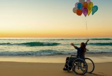 Woman in a wheelchair with her arms in the air on a beach at sunset holding balloons