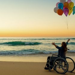 Woman in a wheelchair with her arms in the air on a beach at sunset holding balloons