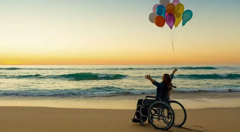 Woman in a wheelchair with her arms in the air on a beach at sunset holding balloons