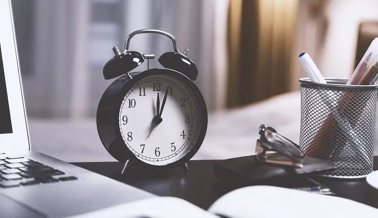 Black clock on a dark wood desk next to a laptop, glasses, pen holder and notebook