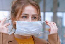 Woman in a beige coat and yellow jumper putting a face mask on