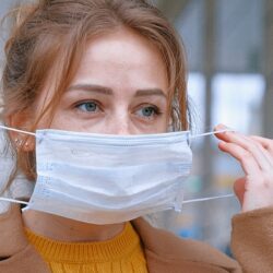 Woman in a beige coat and yellow jumper putting a face mask on