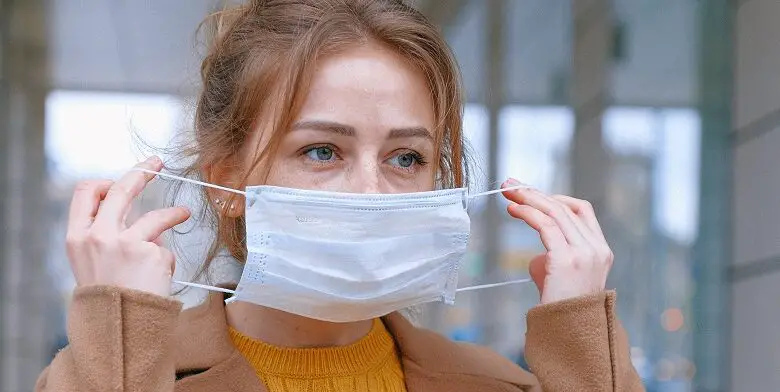 Woman in a beige coat and yellow jumper putting a face mask on