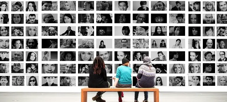 Three people sat on a bench in front of a wall of black and white portraits of people