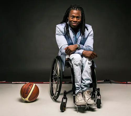 Ade Adepitan sat in his wheelchair in a blue suit jacket and white trousers on a black background with a basketball on the floor next to him