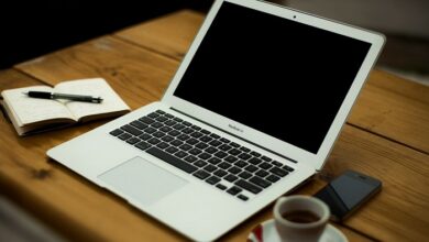 Laptop open on a wooden table with a coffee cup, mobile phone and pad and pen