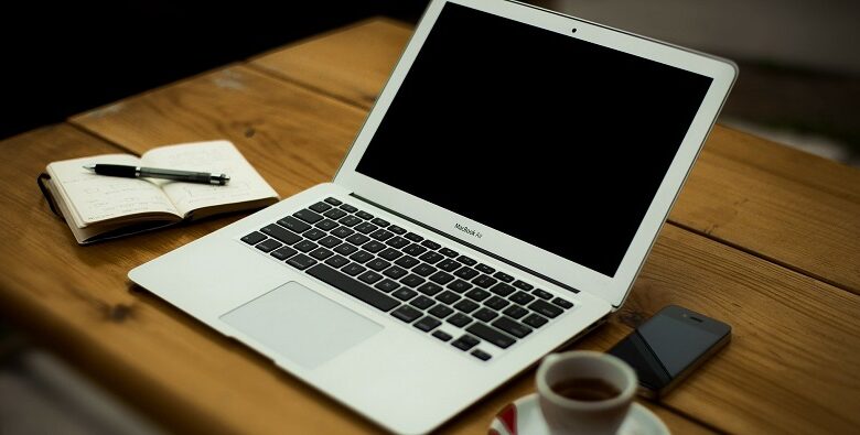 Laptop open on a wooden table with a coffee cup, mobile phone and pad and pen