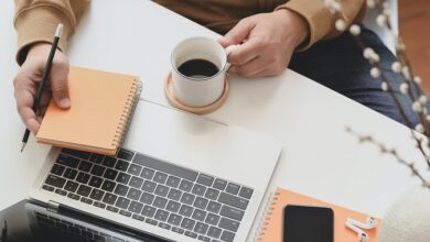 Man with a cup of coffee sat at a desk with a laptop and notebook