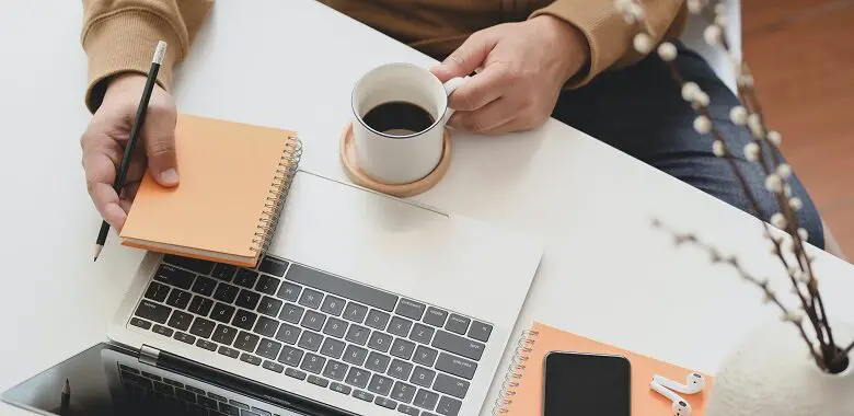 Man with a cup of coffee sat at a desk with a laptop and notebook