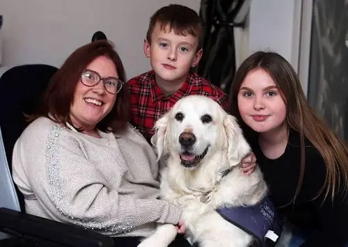 Tara in a white jumper sat in her wheelchair with her two teenage children and assistance dog
