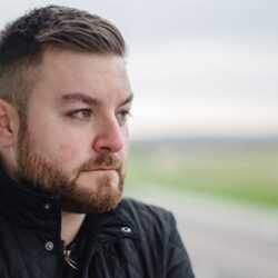 Alex Brooker in a black jacket viewed from the side looking into the distance