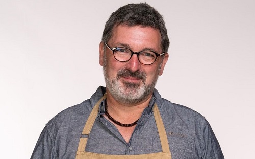 Marc Elliot from the Bake Off in a grey shirt and brown apron with a short beard wearing glasses