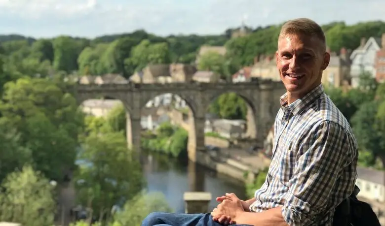 Steve Brown in a checked shirt and jeans sat in his wheelchair with a arched bridge in the background