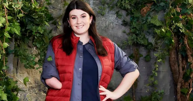 Hollie Arnold in a red puffer jacket and blue tops with her limb difference showing standing in front of a wall covered in leaves ready for I'm a Celebrity 2020
