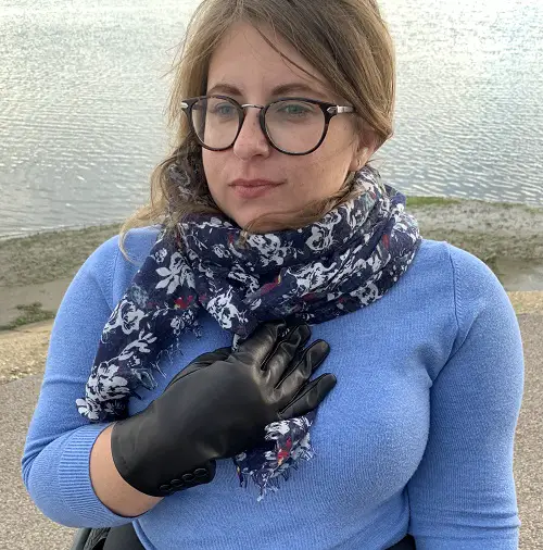 Carina in her wheelchair on a seafront wearing a blue jumper with a black scarf and Hands of Warriors black gloves
