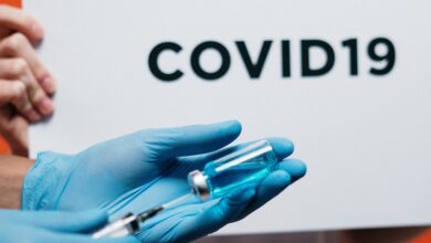 Vaccine being put into a syringe being held in someone's hand wearing blue gloves and a white lab coat in front of a Covid-19 sign