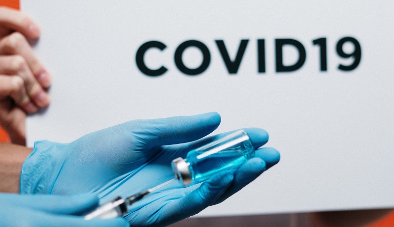Vaccine being put into a syringe being held in someone's hand wearing blue gloves and a white lab coat in front of a Covid-19 sign
