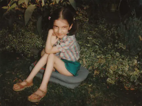Disabled blogger Raya AlJadir as a child sat on cushions on the floor in the garden in front of a bush with her hair in bunches