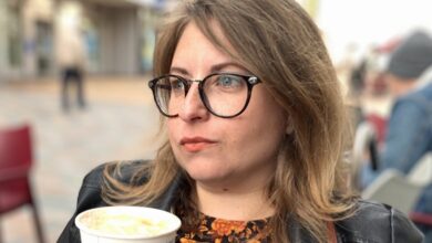 Disabled entrepreneur Carina holding a cup of coffee sitting outsied a cafe looking into the distance wearing glasses