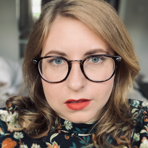 Headshot of disabled entrepreneur Carina with light brown hair wearing brown-rimmed glasses, red lipstick and a black dress with orange and green flowers