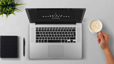 Laptop viewed from above on a table with a man holding a coffee mug and a pad and pen next to it
