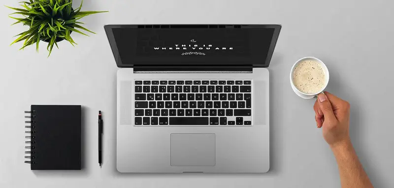 Laptop viewed from above on a table with a man holding a coffee mug and a pad and pen next to it