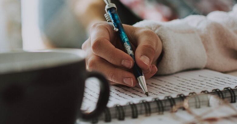 Woman's hand holding a blue pen writing on a pan with a black mug and glasses next to the pad on the table