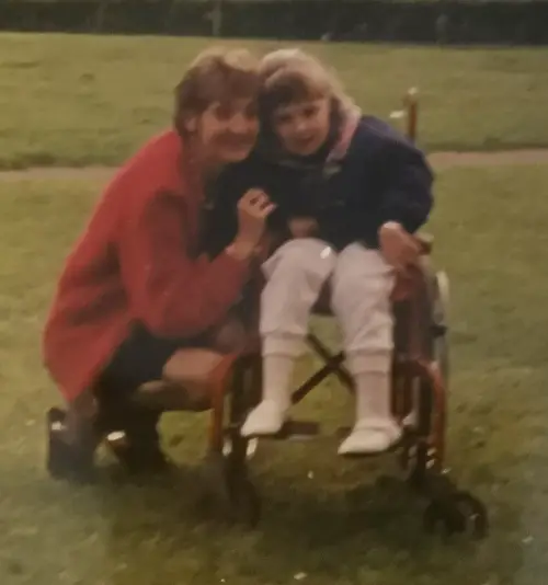 Danielle as a child in a red manual wheelchair outside in a park with her mum knelt next to her