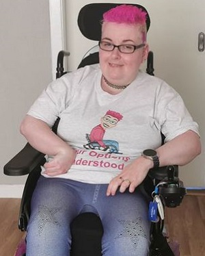 Danielle with pink hair and glasses in her wheelchair wearing a white T-shirt with her logo for Y.O.U and blue jeans with diamante stars on