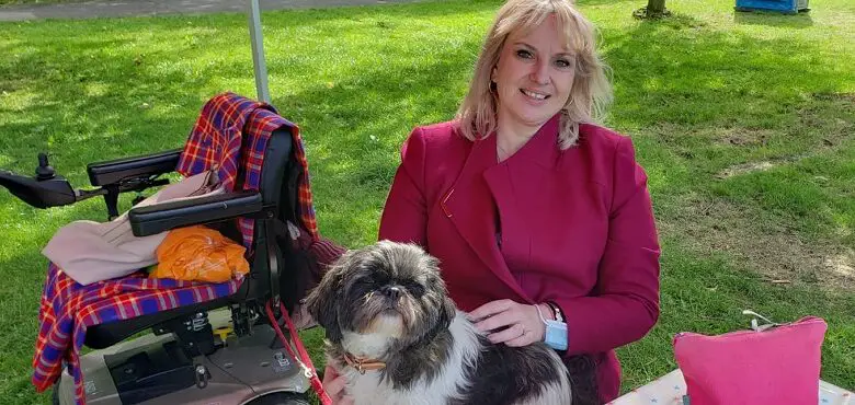 Emma Shepherd wearing a pink coat sat on a bench in a park with her dog on her lap and her mobility scooter behind her