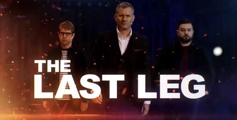 Three men stand in front of a dark background with orange and blue tones and floating sparks. The man in the center wears a suit, while the men on either side wear casual clothing. The text "The Last Leg" is prominently displayed in the foreground. 