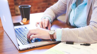 Woman sat at a table working on her laptop with an open notebook and pen next to her